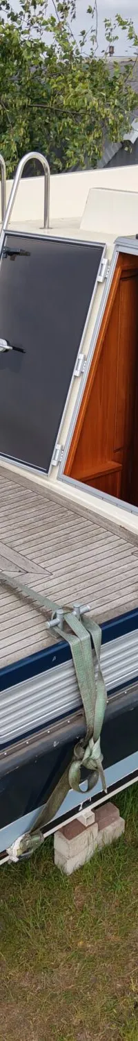 Een blauw-witte boot met een houten dek staat op een trailer in het gras, met bomen en gebouwen op de achtergrond.