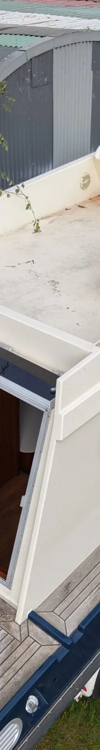 Een crèmekleurige boot met een blauwe romp staat op een trailer op een grasveld, met een open ingang aan de achterkant.