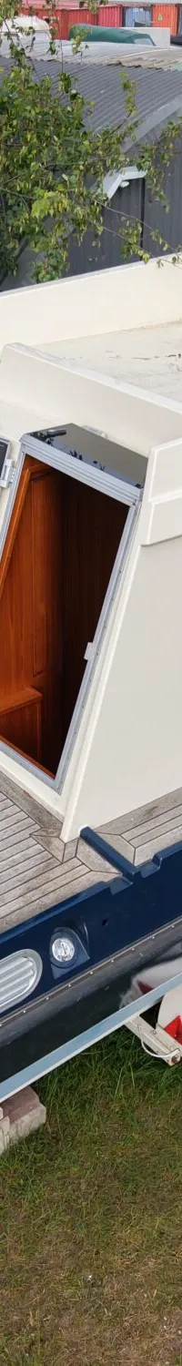 Een blauw-witte boot staat op een trailer in het gras, met een open kajuitdeur en een ladder aan de achterkant.