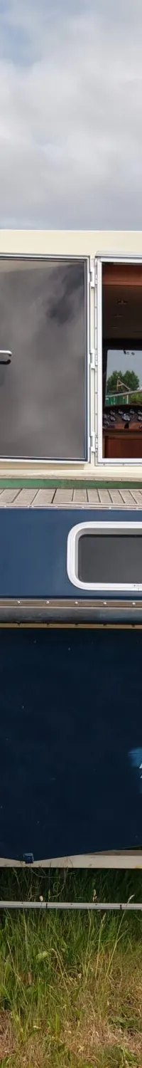 Een blauwe boot met een open kajuit en twee ramen aan de achterzijde staat op een trailer in het gras.