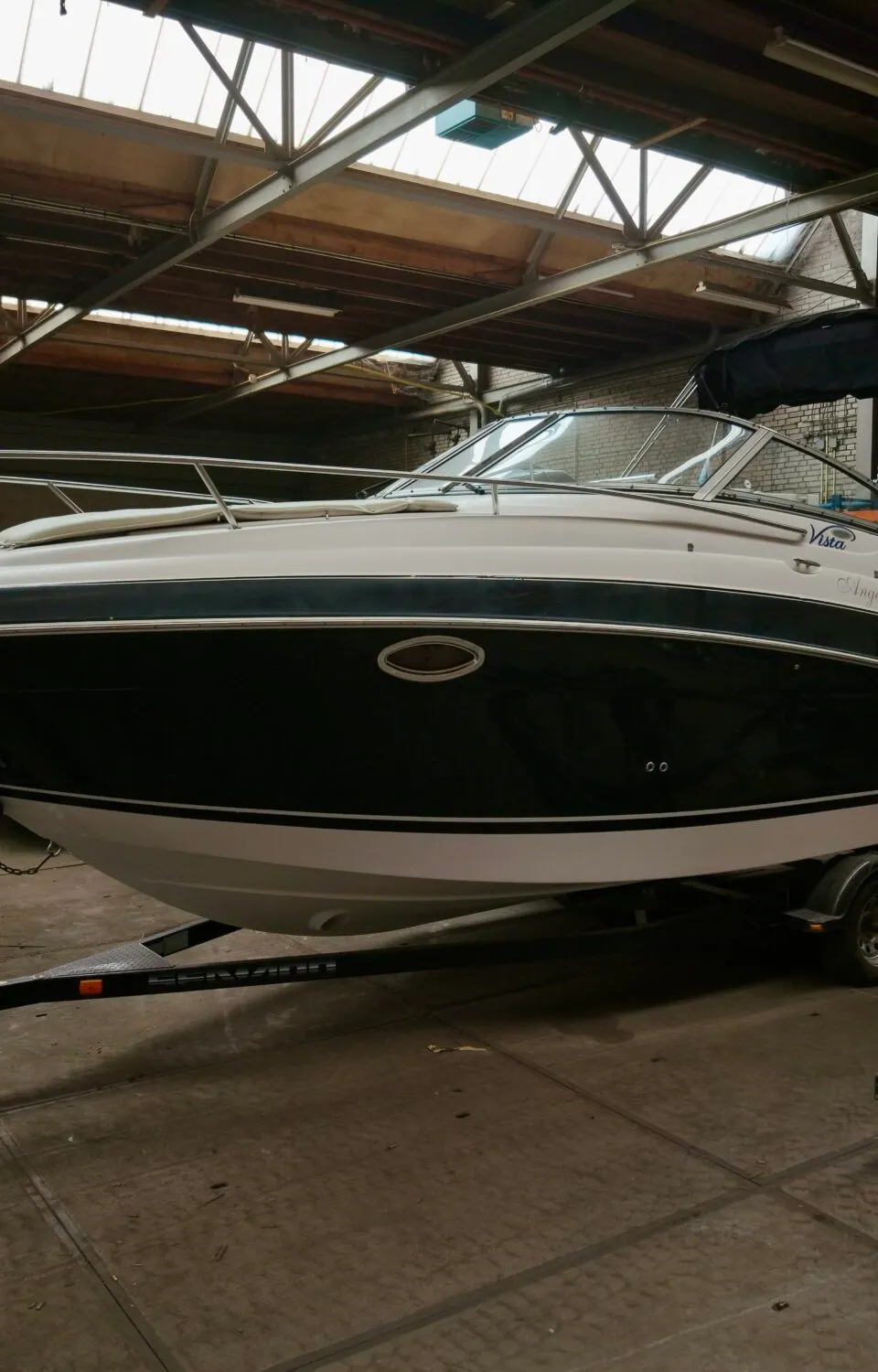 Een witte en donkerblauwe boot staat op een trailer in een loods met een Nederlands vlaggetje aan de boeg.