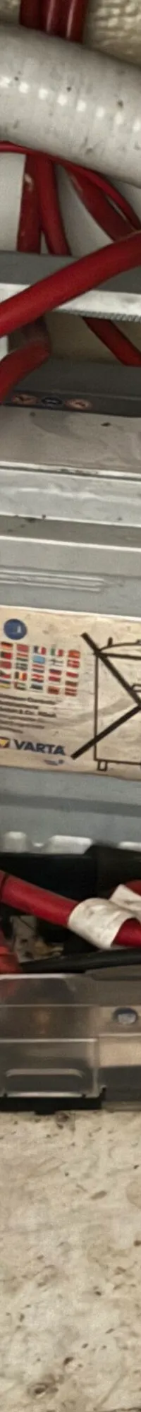 Een grijze Varta-accu met rode en zwarte kabels, een zekeringhouder en diverse leidingen in een rommelige ruimte.