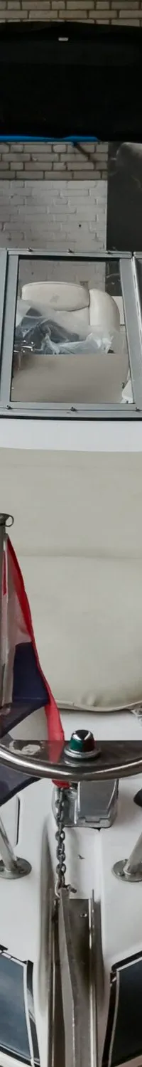 Een witte boot met een zwarte streep en een Nederlandse vlag staat in een loods, omringd door trailers en stellingen.