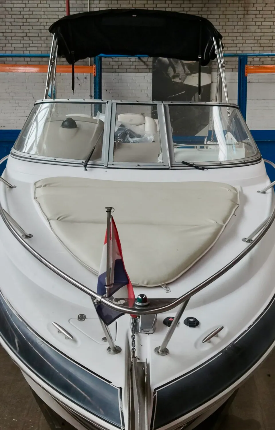 Een witte boot met een zwarte streep en een Nederlandse vlag staat in een loods, omringd door trailers en stellingen.