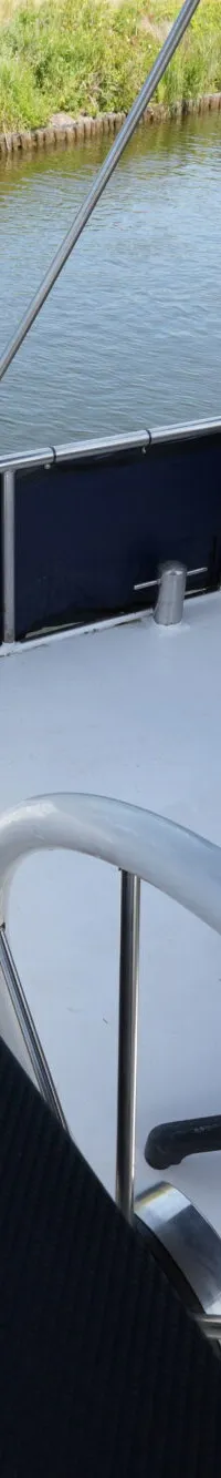 Een stuurstand met een bruine stoel, een chroom gashendel en een stuurwiel op een boot, met water en groen in de achtergrond.