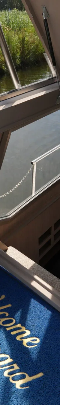 Een blauwe "Welcome Aboard" mat ligt voor een open houten kajuitdeur, met een deel van het stuur aan de rechterkant.