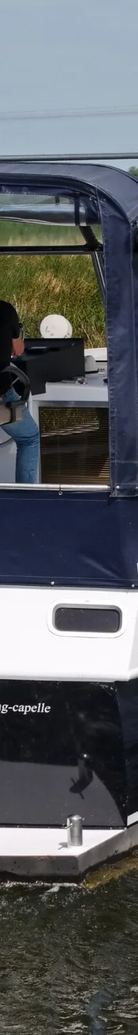 Een blauw-witte motorboot met een blauwe kap vaart op een kanaal, met een persoon aan het roer en een Nederlandse vlag…