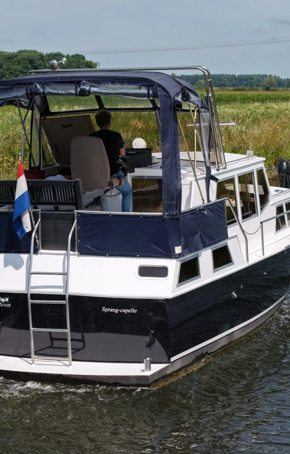 Een blauw-witte motorboot met een blauwe kap vaart op een kanaal, met een persoon aan het roer en een Nederlandse vlag…