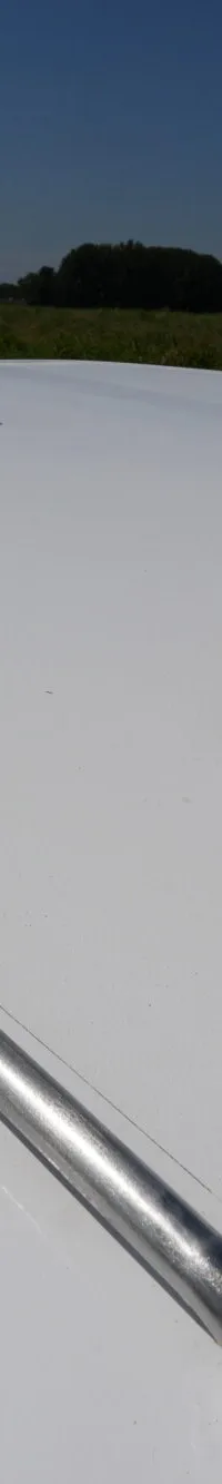 De witte boeg van een boot met een zilverkleurige reling vaart op een kanaal met groene oevers onder een blauwe lucht.