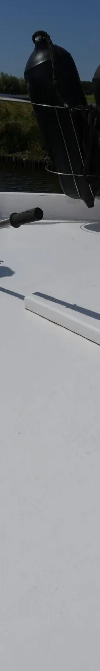 De witte boeg van een boot met drie zwarte stootwillen, een ankerlier en een luik, vaart langs een groene oever.