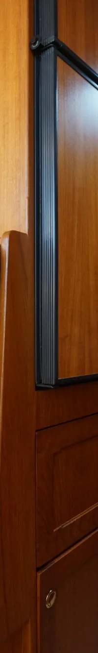 Een kajuit met houten wanden, een bruine bank met kussens, een raam, klokken en een doorgang naar een slaapruimte.