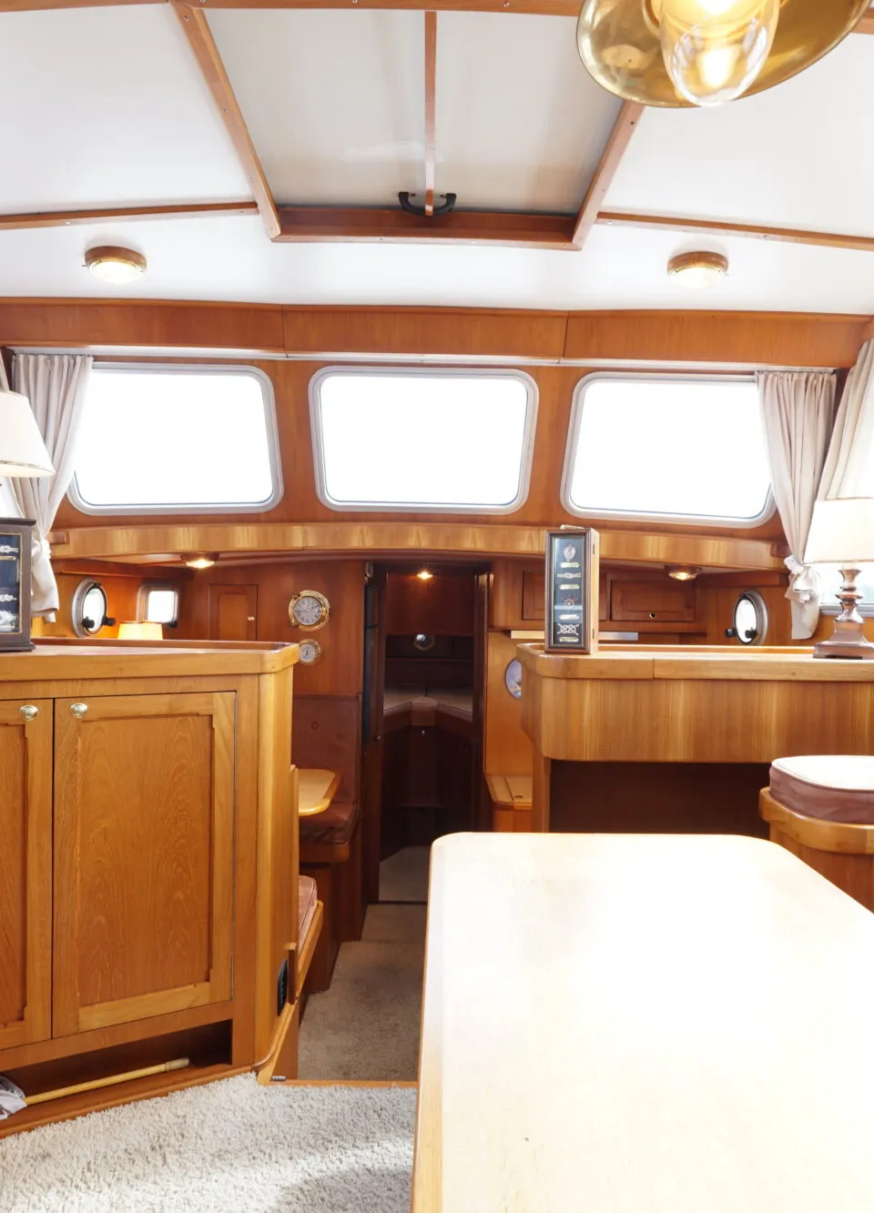 De foto toont het interieur van een boot met houten lambrisering, ramen met gordijnen, een tafel en kussens.