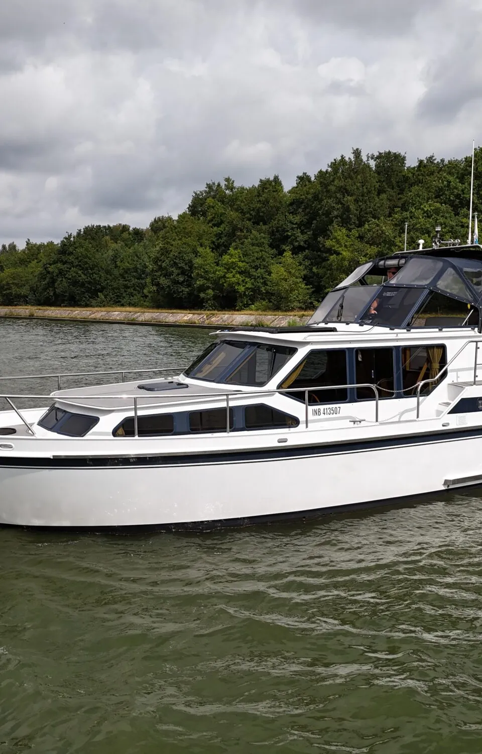 Een witte motorboot met de naam "HYGGE" vaart op een waterweg met bomen langs de oever onder een bewolkte lucht.