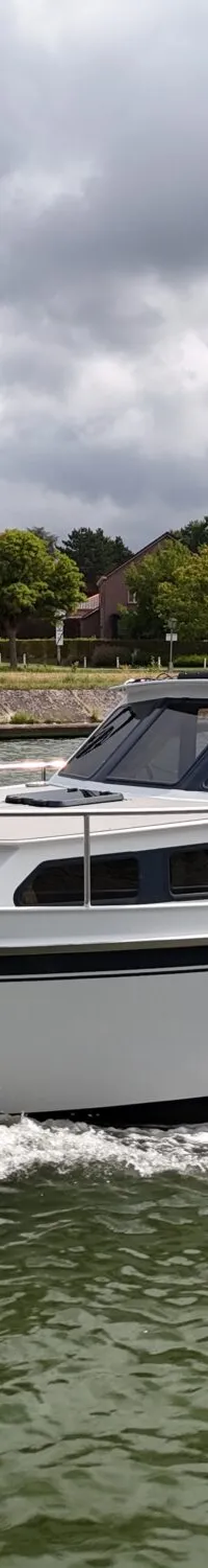 Een witte motorboot met zwarte strepen vaart op een waterweg, met huizen en bomen op de achtergrond onder een bewolkte lucht.