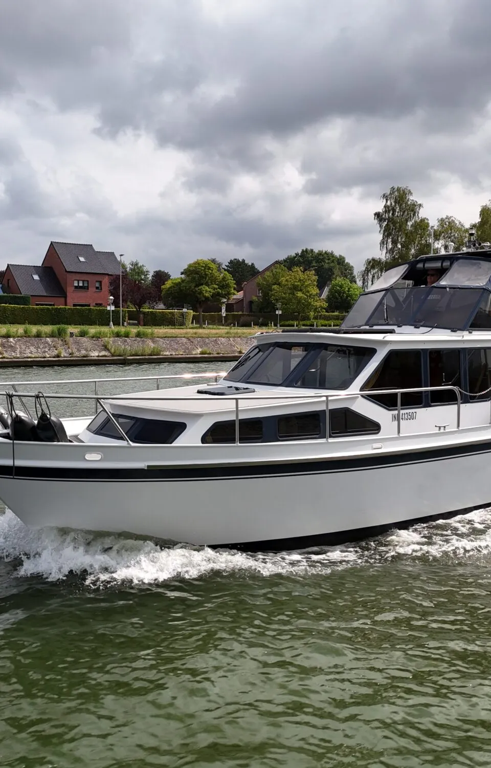 Een witte motorboot met zwarte strepen vaart op een waterweg, met huizen en bomen op de achtergrond onder een bewolkte lucht.