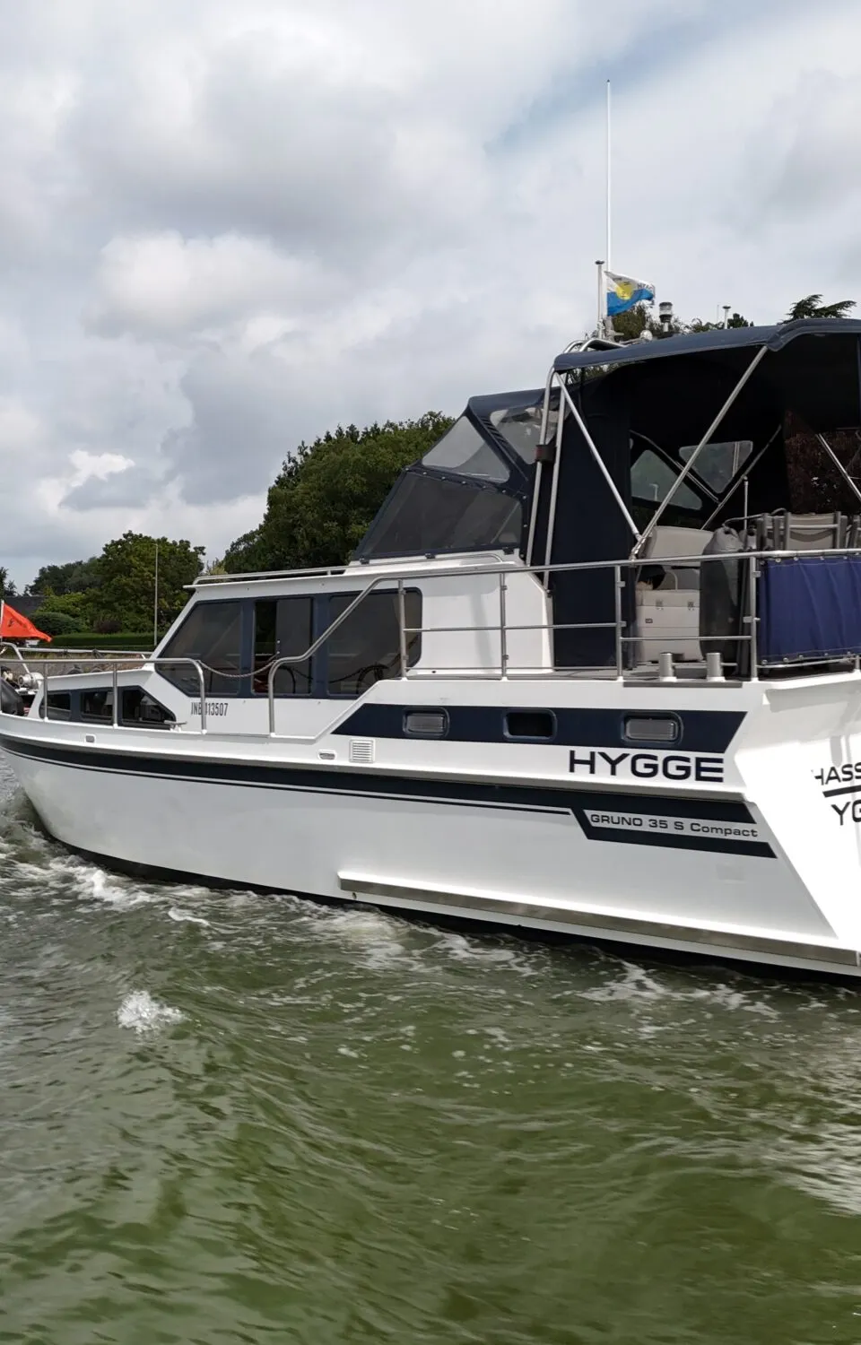 Een witte motorboot met donkerblauwe accenten vaart op een waterweg, met een Belgische vlag aan de achtersteven.
