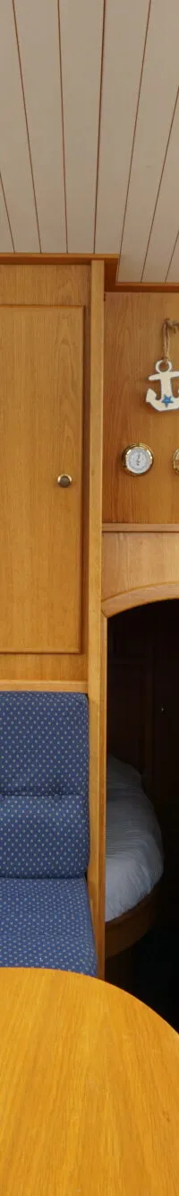 De foto toont het interieur van een boot met een blauwe bank, houten tafel, trap en ramen met gordijnen.