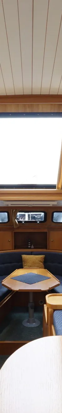 De foto toont het interieur van een boot met een stuurwiel, houten afwerking, blauwe banken en ramen met gordijnen.