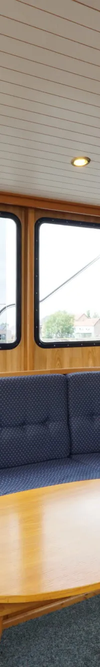 De foto toont een interieur van een boot met een blauwe hoekbank, een houten tafel en ramen met uitzicht naar buiten.