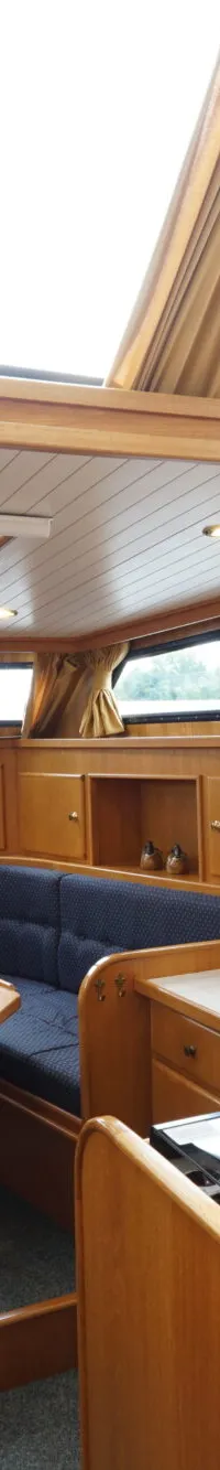 De foto toont het interieur van een boot met houten afwerking, een zithoek met blauwe kussens, een tafel, een keukenblok en…