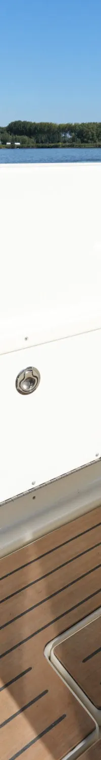 De achterzijde van een boot met een wit compartiment, een bruine zitting en een houten dek, varend op water met industrie…