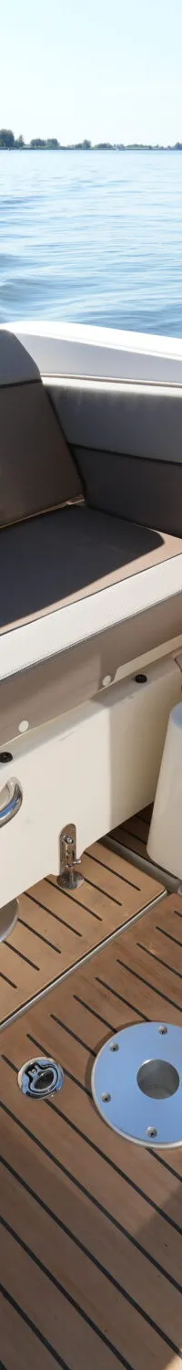 De cockpit van een boot met houten vloer en beige/bruine zitkussens is zichtbaar, met water en landschap op de achtergrond.