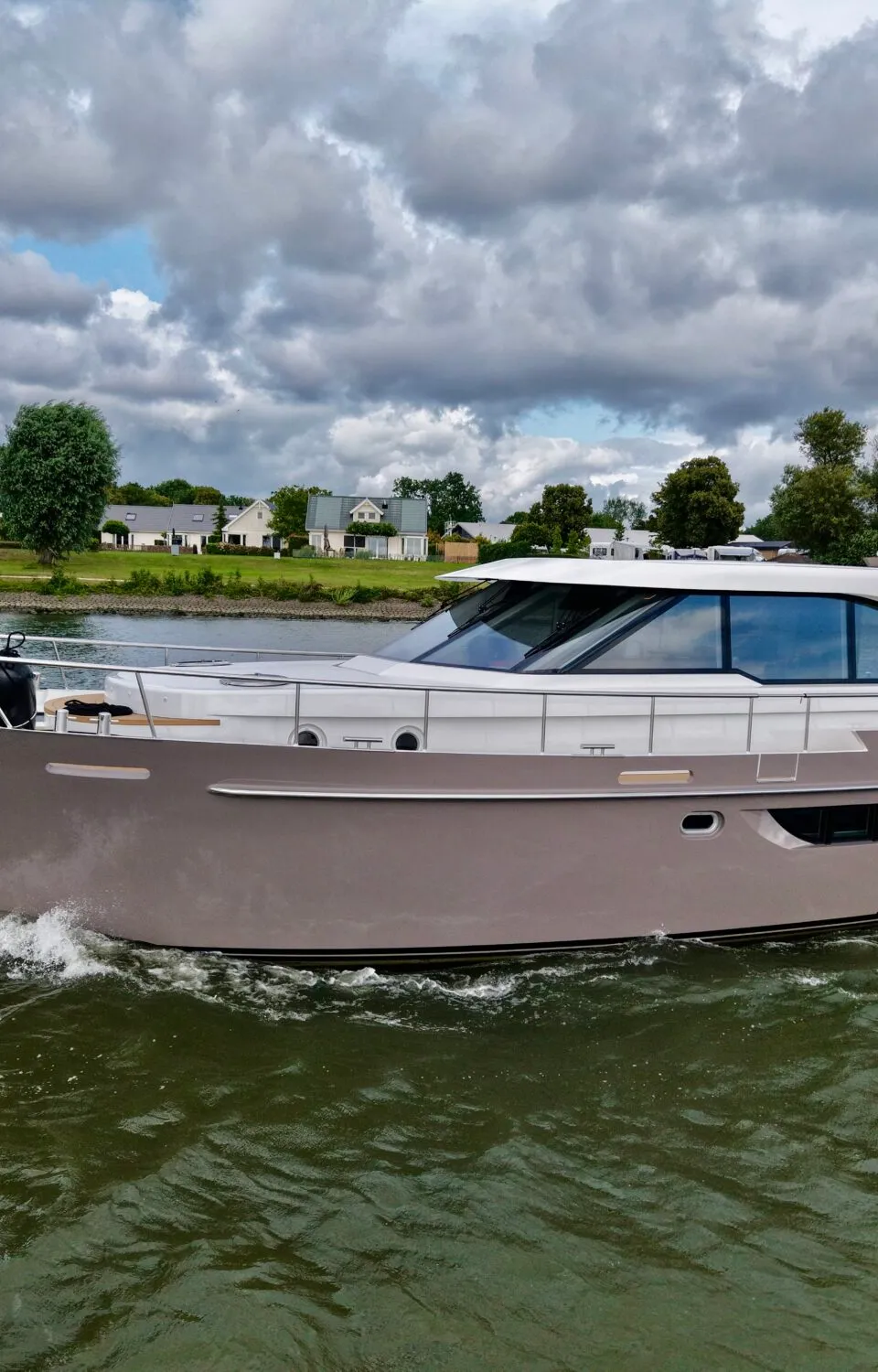 Een beige met witte motorboot genaamd 'SRIJN VERIUS' vaart op water langs een oever met huizen en bomen onder een bewolkte…