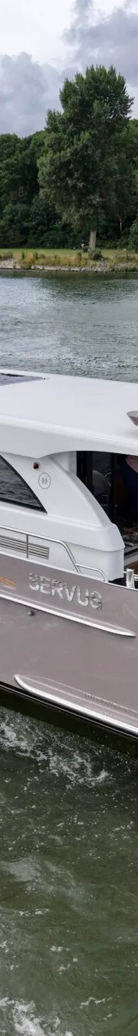 Een varende motorjacht met de naam "SERVUS" op de romp, toont een Nederlandse vlag aan de spiegel en creëert een hekgolf.