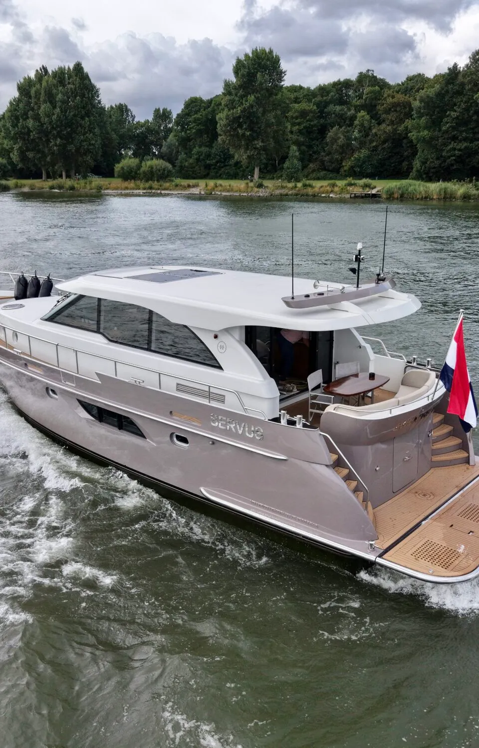 Een varende motorjacht met de naam "SERVUS" op de romp, toont een Nederlandse vlag aan de spiegel en creëert een hekgolf.