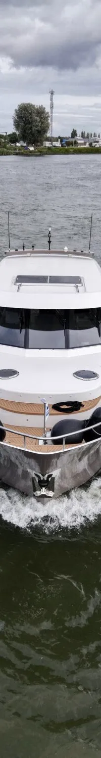 Een witte motorjacht vaart op een waterweg met een lichte boeggolf, met een dorp en een vrachtschip op de achtergrond.