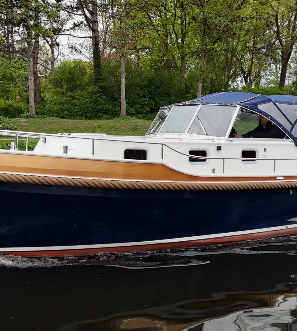 Een blauw-witte motorboot met een bruine stootrand en een blauwe kap vaart op een waterweg langs een groene oever.