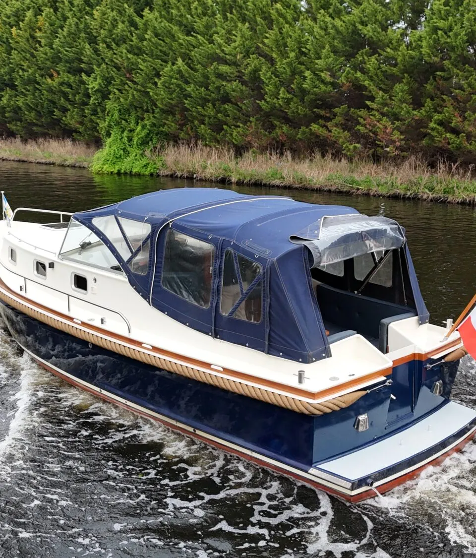 Een blauw-witte motorboot met een blauwe kap vaart over een waterweg met een vlag aan de achtersteven.