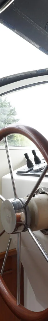 Een houten stuurwiel, een blauwe stoel en een dashboard met bedieningselementen zijn zichtbaar in de stuurhut van een boot.