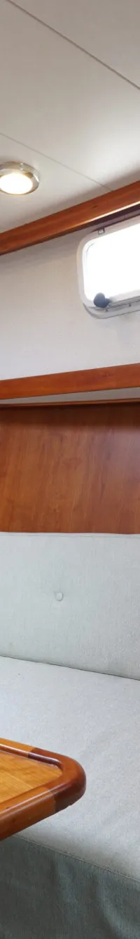 De foto toont een kajuit met een houten tafel, een bank met lichtgroene kussens en twee sierkussens.