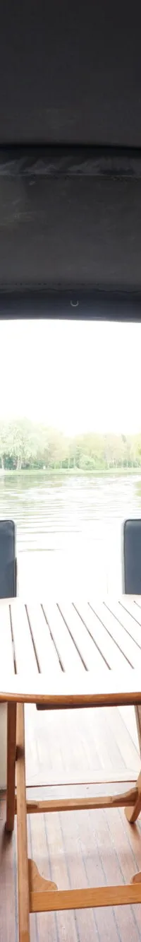 De foto toont het interieur van een boot met een houten tafel en blauwe kussens, omgeven door een zwarte kap.