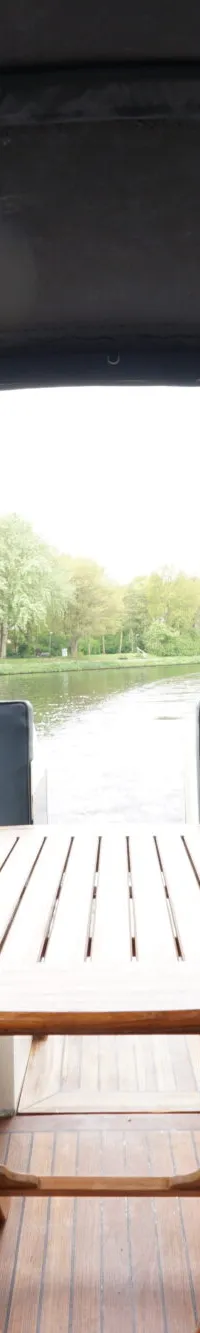 Een interieur van een boot met twee blauwe banken, een houten tafel en uitzicht op water en bomen.