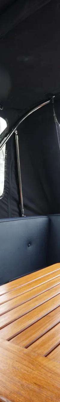 Een interieur van een boot met een houten tafel, donkerblauwe kussens en een uitzicht op water en groen.