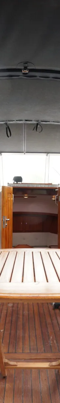 De foto toont het interieur van een boot met een houten vloer, twee blauwe banken, een houten tafel en twee stuurstoelen.