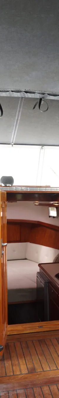 De foto toont het interieur van een boot met twee blauwe stoelen, een houten stuurwiel en een open kajuitdeur.
