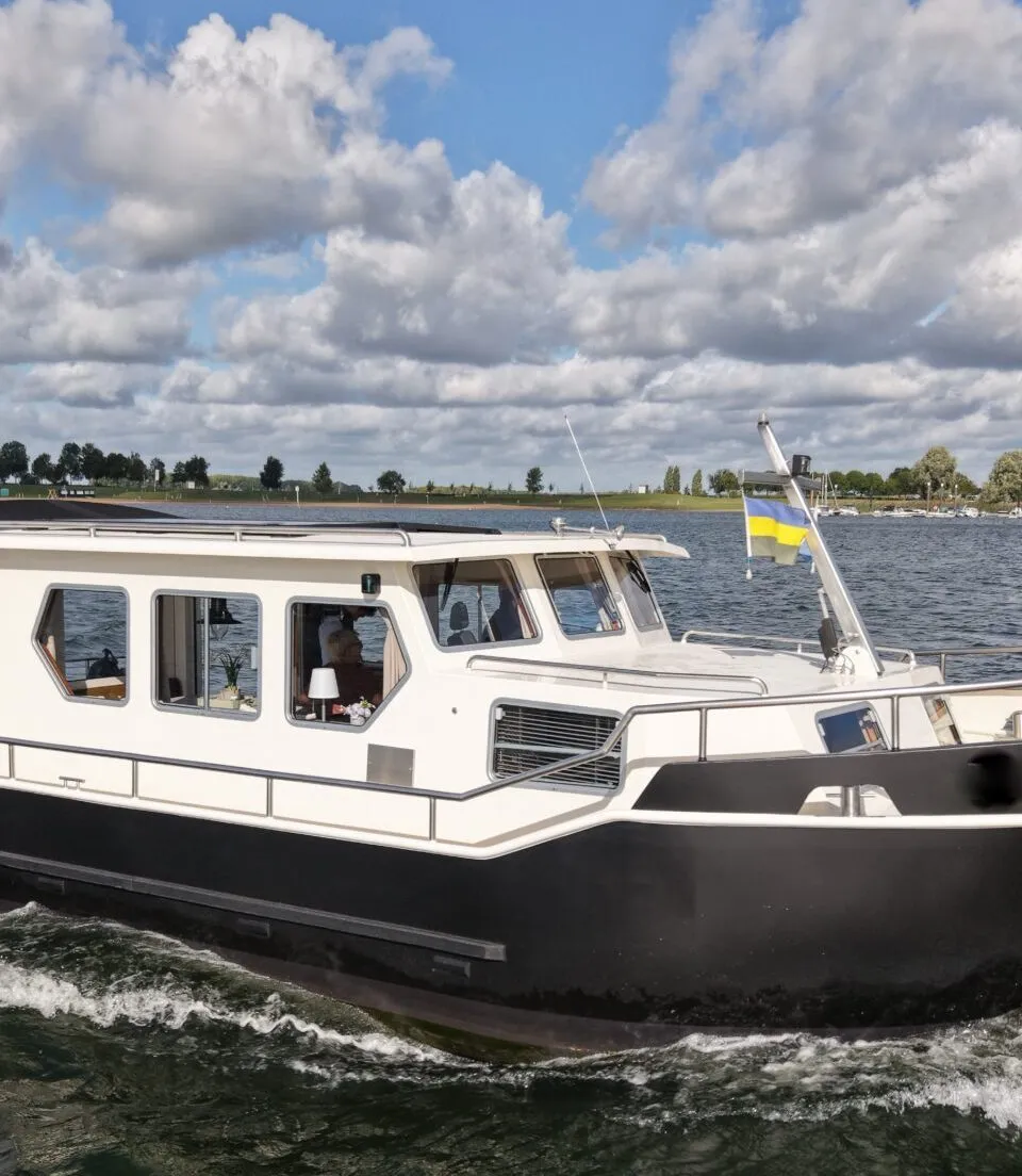 Een witte en zwarte motorboot vaart op het water onder een bewolkte hemel, met land en bomen op de achtergrond.