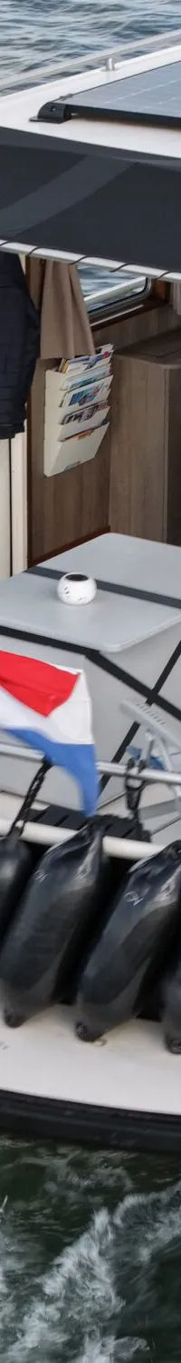 Een witte boot met zwarte romp vaart op het water, met zonnepanelen op het dak en een Nederlandse vlag aan de achtersteven.