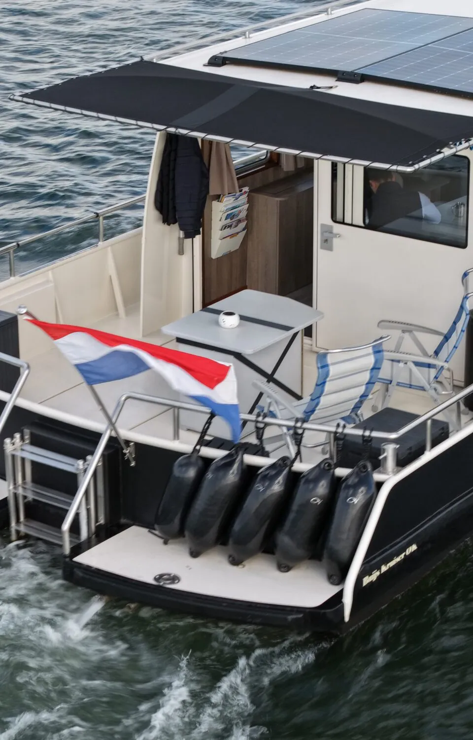 Een witte boot met zwarte romp vaart op het water, met zonnepanelen op het dak en een Nederlandse vlag aan de achtersteven.