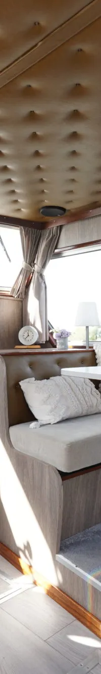 Een interieur van een boot met een stuurstoel, een eettafel met banken, ramen en een gestoffeerd plafond.
