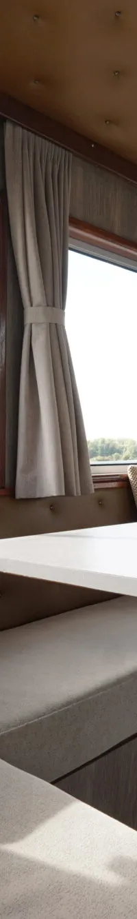 Een interieur van een boot met een tafel, banken met kussens, ramen met uitzicht en een hanglamp.