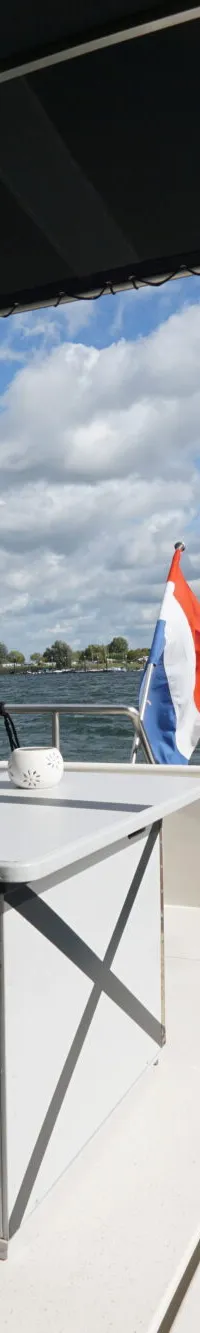 Een bootdek met twee stoelen, een tafel, een opbergkist en een Nederlandse vlag, met water en landschap op de achtergrond.
