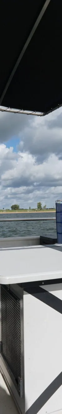 Een dek van een boot met een tafel, twee stoelen, een Nederlandse vlag en uitzicht op water en landschap.