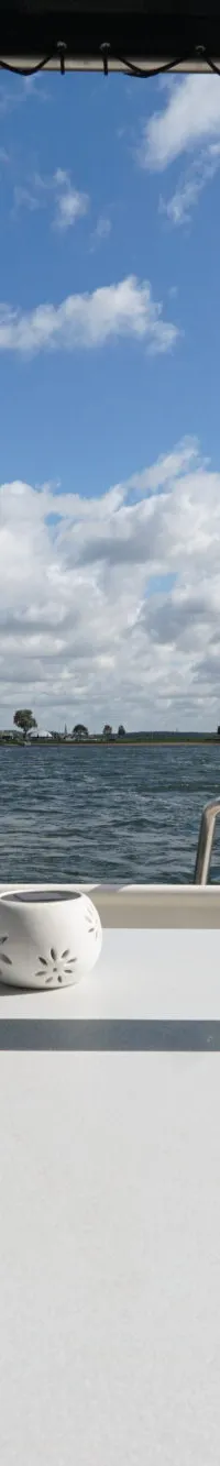 Een bootdek met een tafel, stoel, vlag en uitzicht op water en landschap onder een bewolkte hemel.
