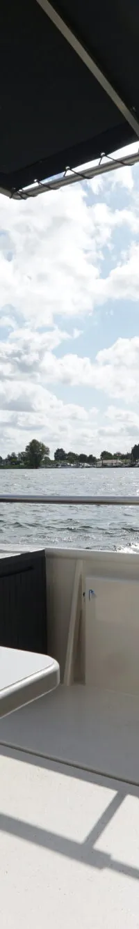 Een bootdek met een tafel en een wit ornament, met uitzicht op water en de horizon met bomen en huizen.
