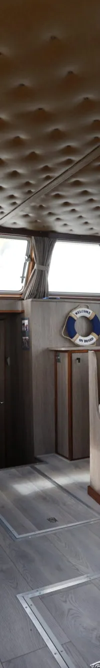 De foto toont het interieur van een boot met een stuurstoel, een tafel met zitbanken en ramen rondom.