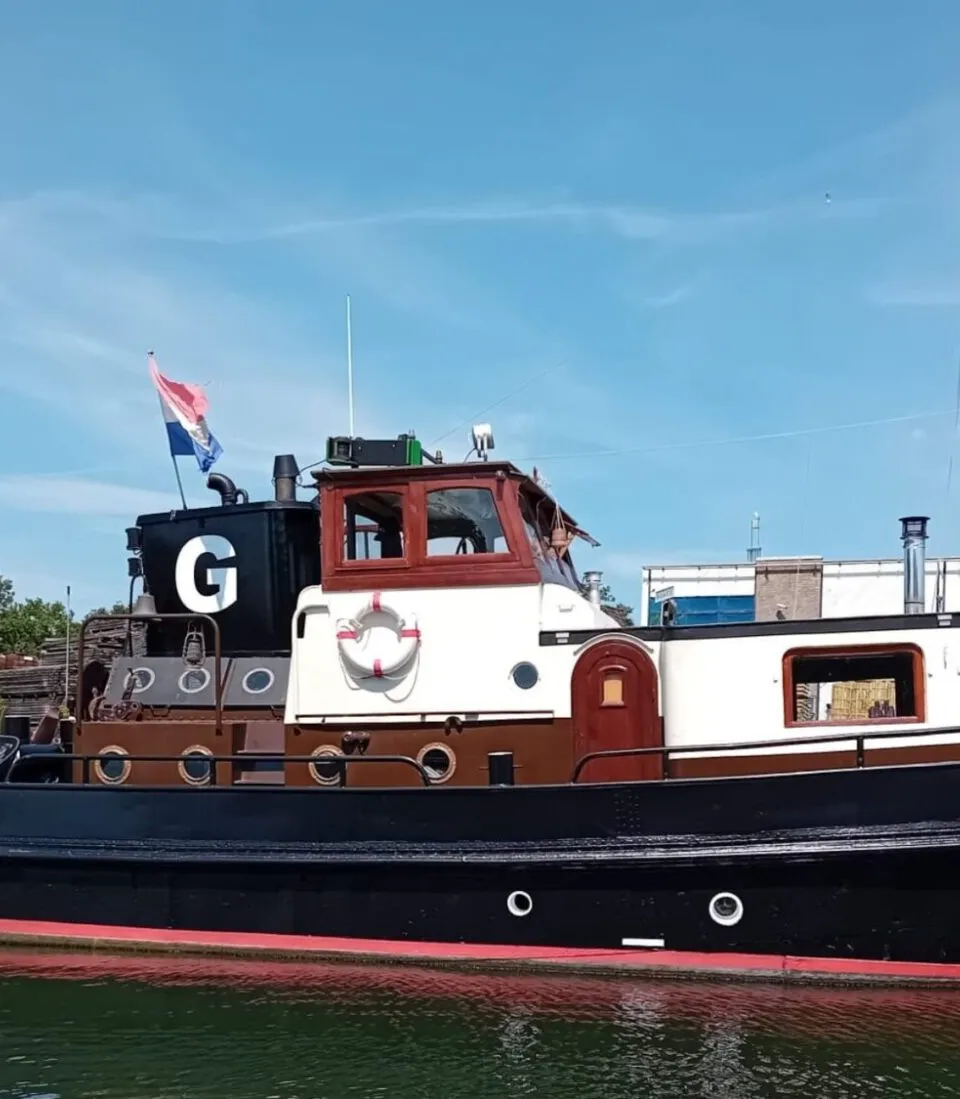 Een zwarte boot met een rode waterlijn en witte opbouw, met de naam "JAN ABELS" op de boeg en een Nederlandse vlag achterop.